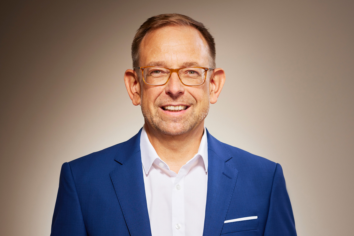 Porträt eines mittelalten Mannes mit kurzem, hellbraunem Haar, Brille und leichtem Bartschatten. Er trägt einen gut sitzenden blauen Anzug mit einem weißen Hemd und hat ein freundliches Lächeln. Der Hintergrund ist in warmen, neutralen Tönen gehalten, und das professionelle Licht betont sein Gesicht gleichmäßig.